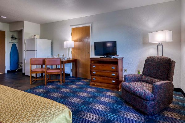 A hotel room features a patterned chair, a TV on a dresser, a small table with two chairs, a refrigerator, and an ironing board in the corner.