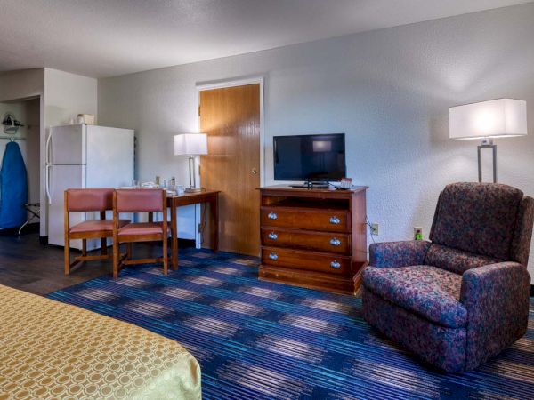 A hotel room features a patterned chair, a TV on a dresser, a small table with two chairs, a refrigerator, and an ironing board in the corner.