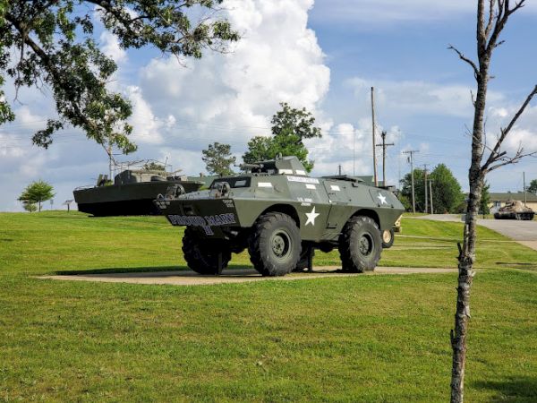The image shows a military vehicle displayed on a grassy field under a partly cloudy sky, with trees and a clear path nearby.