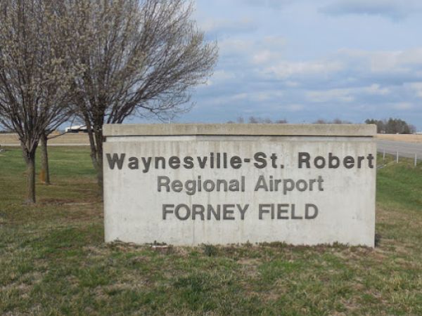A sign for Waynesville-St. Robert Regional Airport, Forney Field, surrounded by grass and trees on a cloudy day.