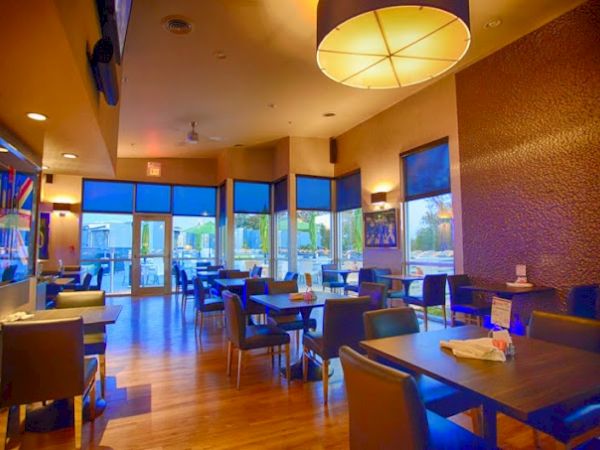 A cozy restaurant interior with modern decor, wooden flooring, and tables set for dining. Large windows show an outdoor seating area.