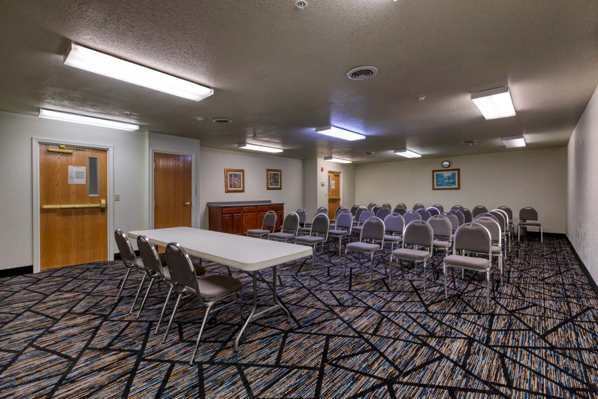 The image shows a conference room with rows of chairs, a table, a patterned carpet, and framed pictures on the walls.