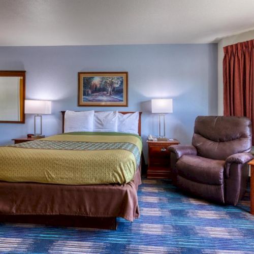 A hotel room with a bed, recliner, lamps, artwork, and a window with curtains. The decor features warm and neutral tones.