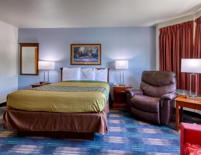 A hotel room with a bed, recliner, lamps, artwork, and a window with curtains. The decor features warm and neutral tones.
