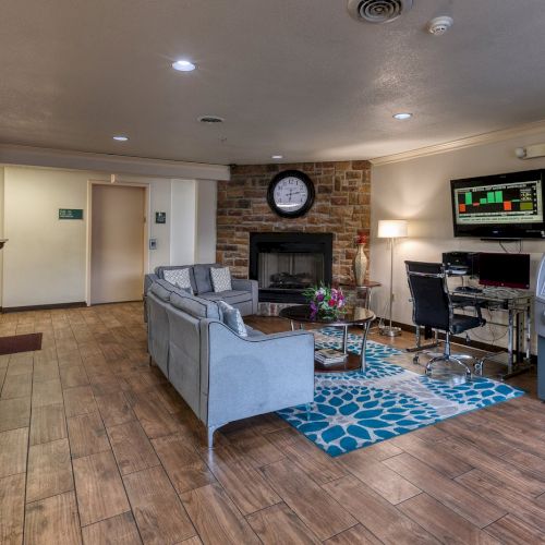 The image shows a cozy lobby area with sofas, a fireplace, clock, TV, computer desk, and an ATM. The floor has a wood-like finish.