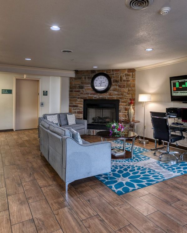 The image shows a cozy lobby area with sofas, a fireplace, clock, TV, computer desk, and an ATM. The floor has a wood-like finish.