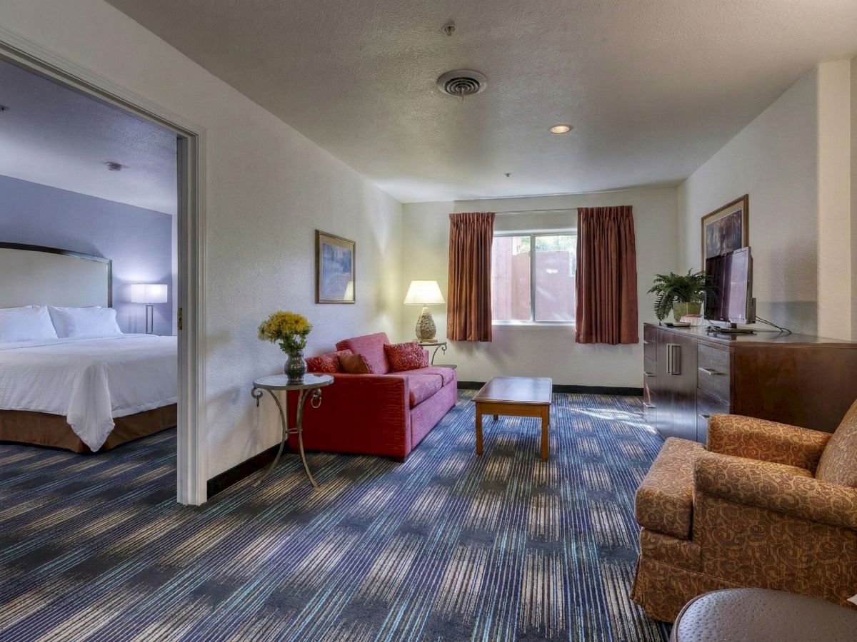 A motel suite with a living area featuring a red sofa, a coffee table, a sofa chair, and a dresser with a TV; doorway leads to a separate bedroom with a king bed.