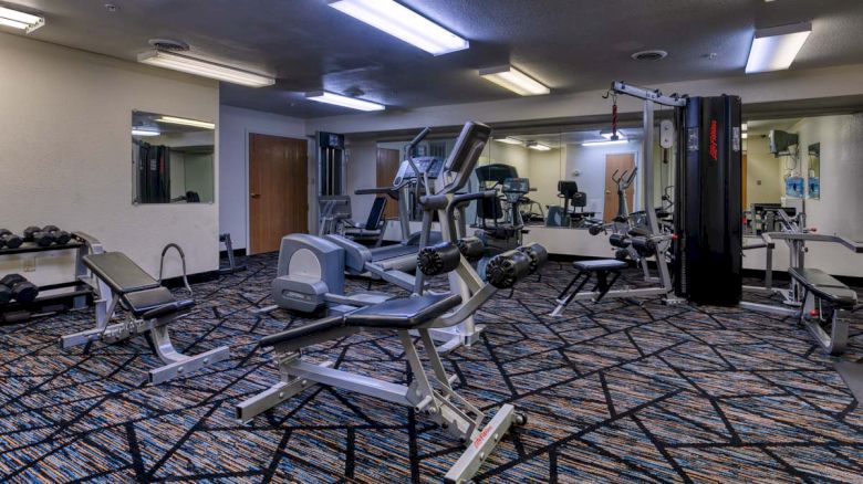 A gym with exercise equipment, mirrors, and carpet flooring, featuring machines like ellipticals and benches.