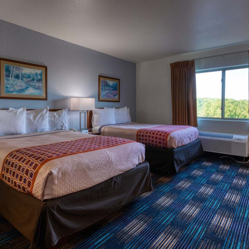 The image shows a hotel room with two double beds, a window with curtains, framed art, bedside tables, and a patterned carpet.