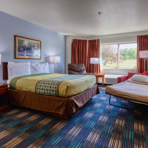 A hotel room with a large bed, nightstands, desk chair, artwork, window, curtains, patterned carpet, and a fold-out couch with bedding.