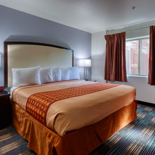 A neatly made hotel room with a large bed, nightstands, lamps, a digital clock showing 6:59, and a window with closed curtains.