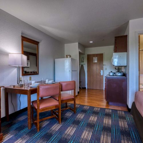 The image shows a hotel room with a TV, table, chairs, kitchenette, and a glimpse of a bathroom with a bathtub and curtain.