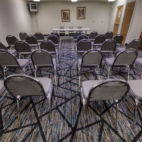 The image shows a conference room with rows of empty chairs facing a table at the front, and a patterned carpet.