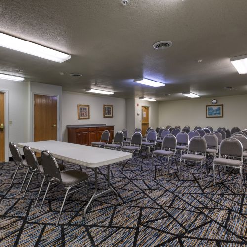 The image shows an empty conference room with rows of chairs, a table, and fluorescent lighting, featuring framed artwork on the walls.