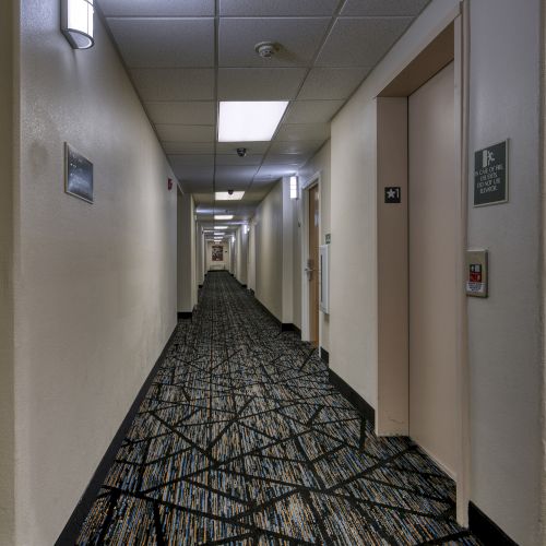 A long, carpeted hotel hallway with doors on both sides, overhead lights, and a small sign with instructions near the elevator.