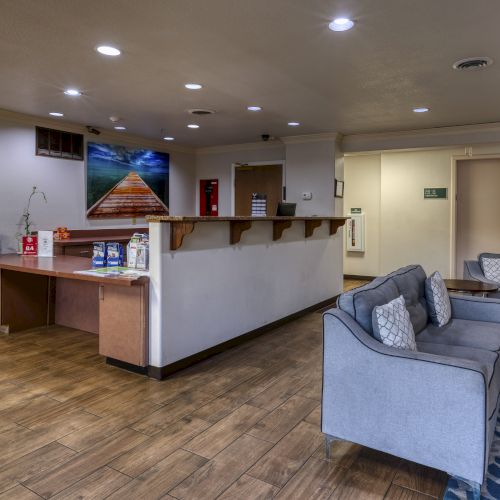 The image shows a cozy lobby with a reception desk, seating area, brochures, artwork, and a patterned rug on a wooden floor.