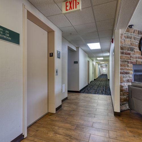 A hotel hallway features an exit sign, elevator, clock on a stone wall, and a sofa. Room direction signs are also visible in the image.