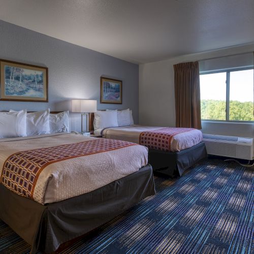 The image shows a hotel room with two double beds, patterned bedding, a window with a view, two framed pictures, and carpeted floor.