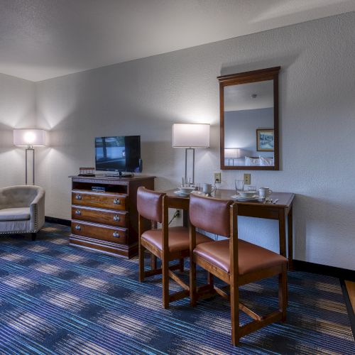 The image shows a hotel room with a TV, a table with chairs, and a fridge. There's a mirror, a lamp, and an armchair nearby.