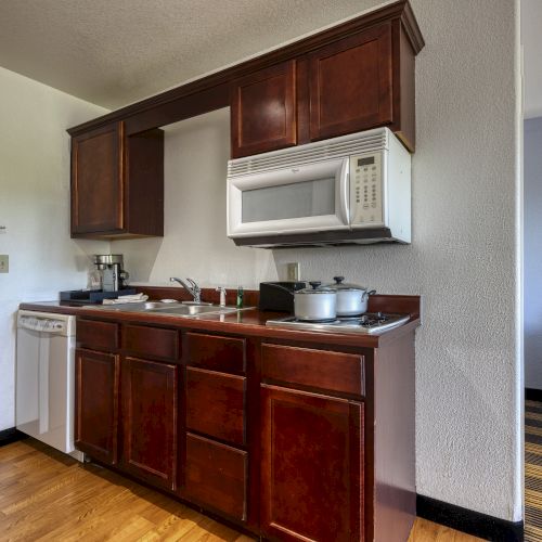 A hotel kitchenette with a microwave, stove, sink, coffee maker, and cabinets. A bed is visible in the adjacent room.