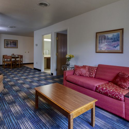 A living room with a red sofa, coffee table, dining area, and artwork on the wall. The space has carpet and open bathroom doors nearby.