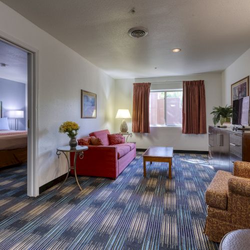 A hotel room with a bed, sitting area, red couch, patterned armchair, TV, and window with curtains, creating a cozy ambiance.