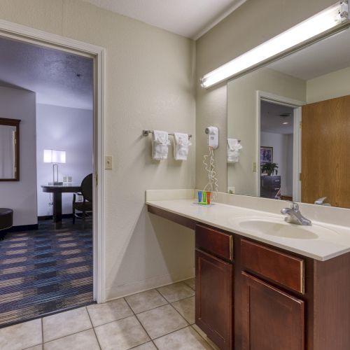 A hotel bathroom with a sink, mirror, wall-mounted hairdryer, and towels; an adjoining room with a table and lamp is visible.