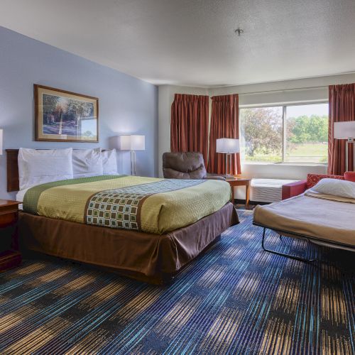 The image shows a hotel room with a bed, a fold-out sofa, lamps, and a window with curtains. A painting is on the wall.