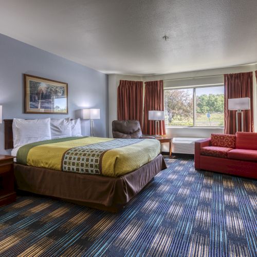A hotel room with a bed, nightstands, lamps, a sofa, a chair, carpet, curtains, and a window with a view outside.