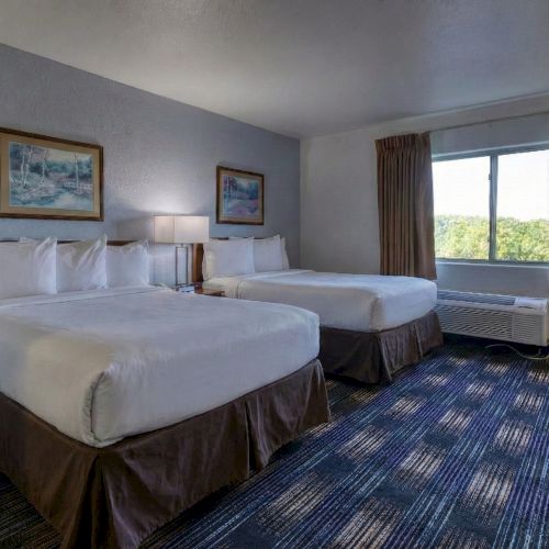 A tidy hotel bedroom with two twin beds, framed artwork, a desk, window with drapes, and soft lighting.