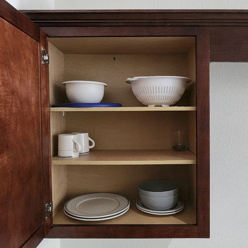 An open kitchen cabinet with plates, bowls, cups, and a colander neatly arranged on two shelves.