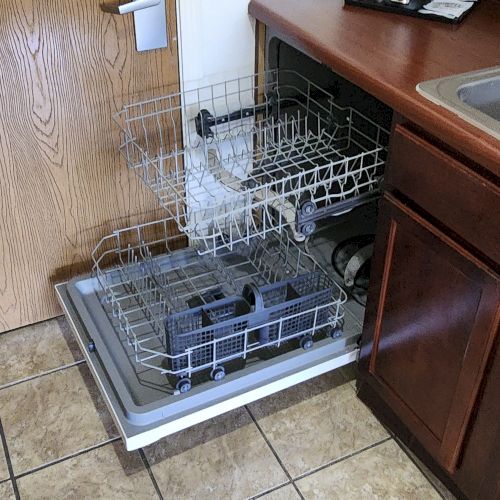 An open dishwasher beside a kitchen sink, with wooden cabinets and a tiled floor.