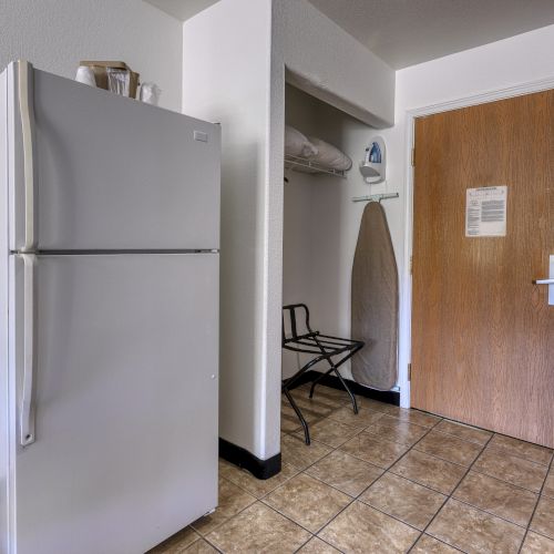 The image shows a room with a refrigerator, an ironing board, a luggage rack, and a door, featuring tiled flooring and minimal furnishings.