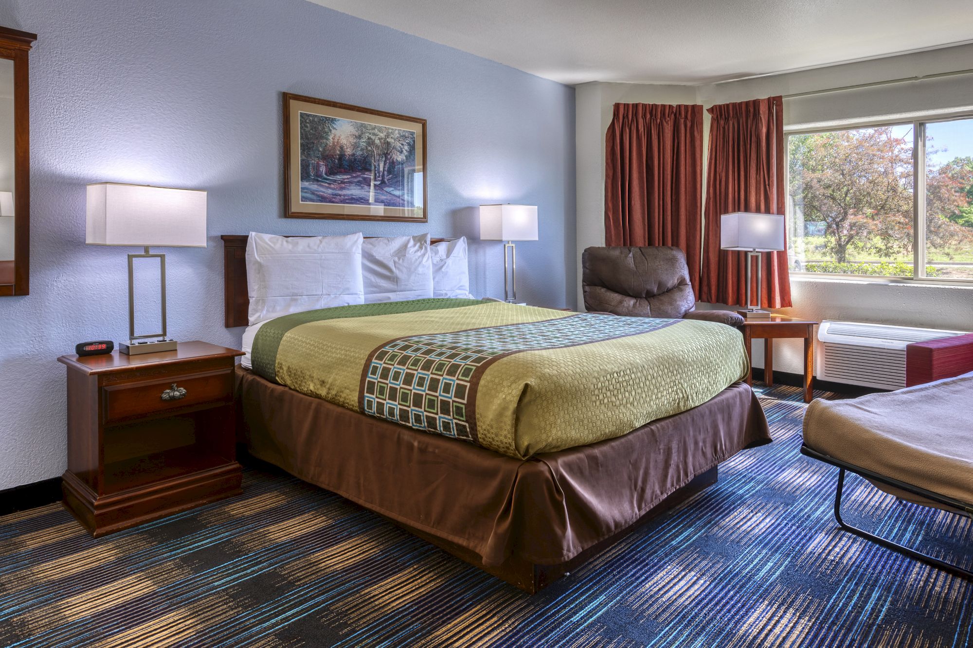 The image shows a hotel room with a bed, two bedside lamps, a chair, artwork, curtains, and a window with a view of trees outside.