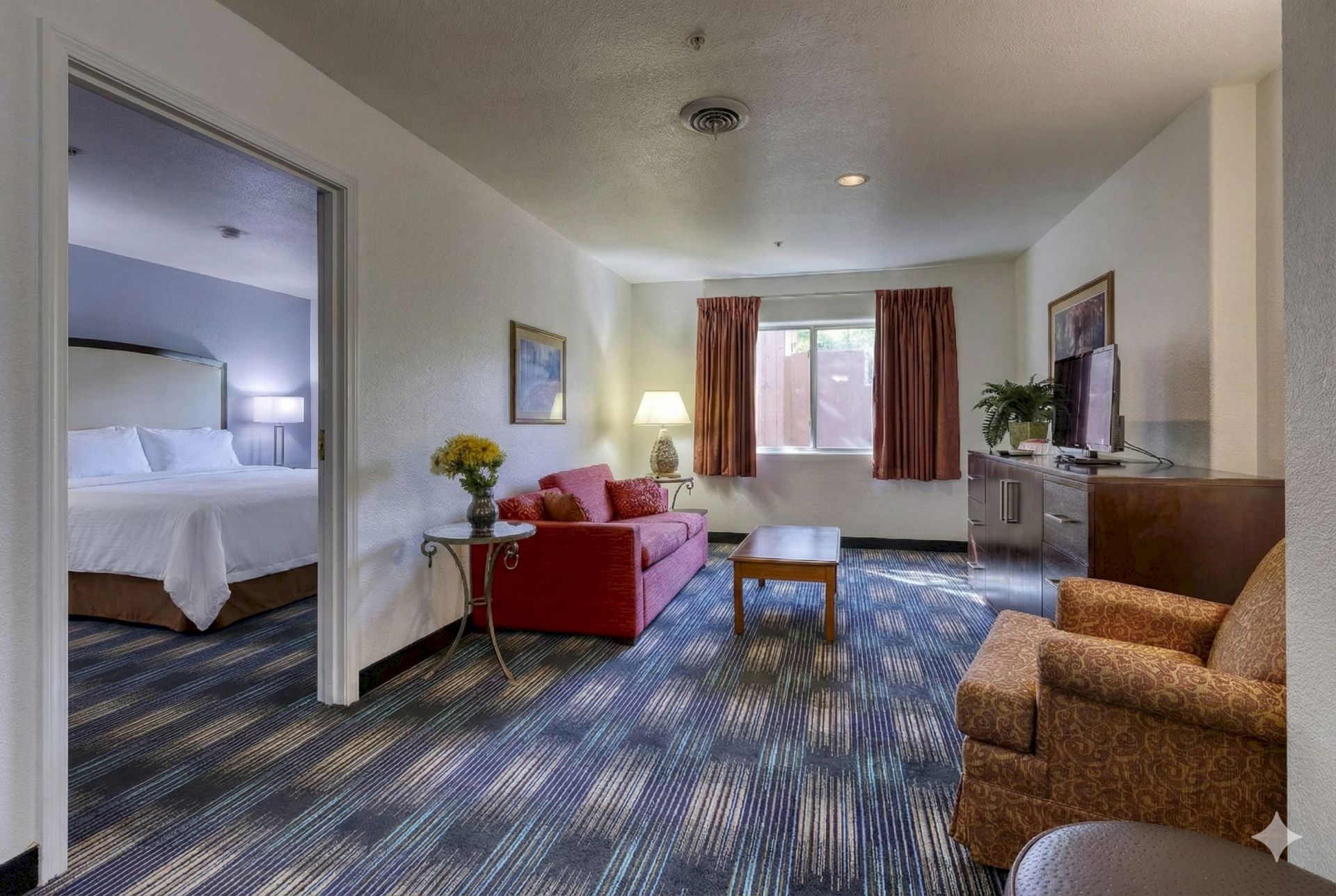 A tidy hotel living room with couch, coffee table, TV stand, and a view into a bedroom with beds.