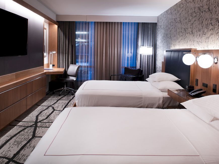 A modern hotel room with two beds, a TV, a desk, and a large window with curtains. The design is sleek and minimalist.