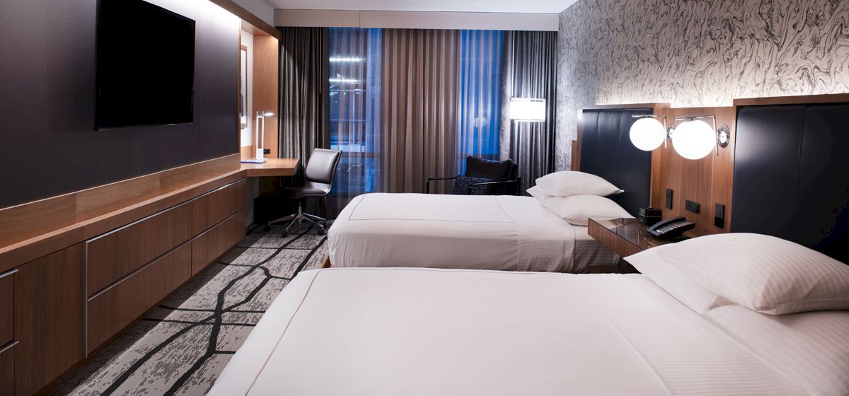 A modern hotel room with two beds, a TV, a desk, and a large window with curtains. The design is sleek and minimalist.