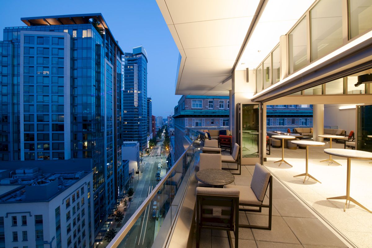 A cityscape at twilight with modern buildings and a balcony featuring tables and chairs overlooking the street scene below.