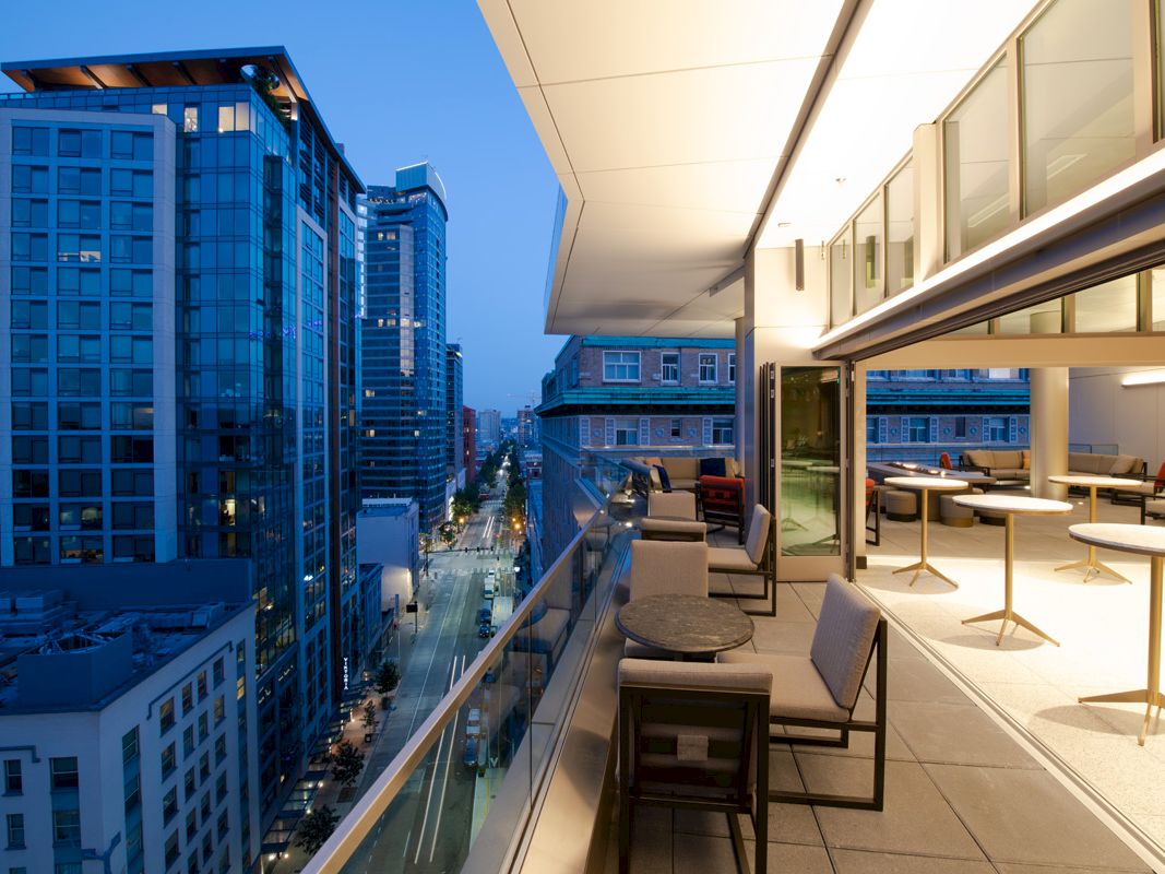 A cityscape at twilight with modern buildings and a balcony featuring tables and chairs overlooking the street scene below.