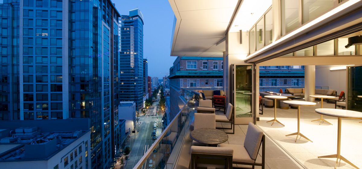A cityscape at twilight with modern buildings and a balcony featuring tables and chairs overlooking the street scene below.