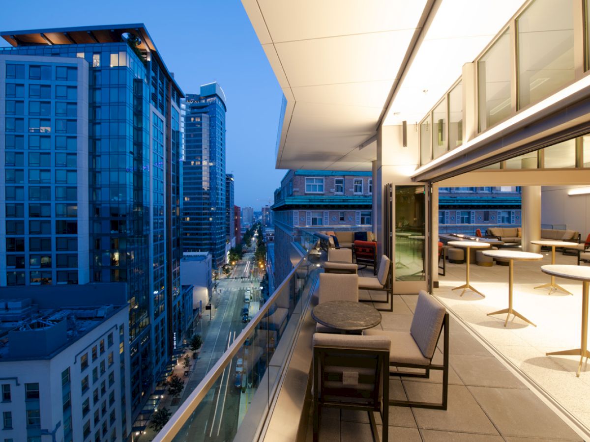 A modern rooftop terrace with seating overlooks a city street lined with tall buildings at dusk.