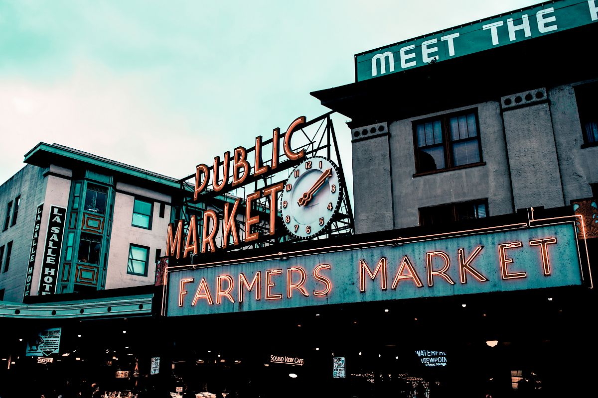 The image shows a neon sign reading "Public Market" and "Farmers Market" with a clock, against a backdrop of buildings under a cloudy sky.