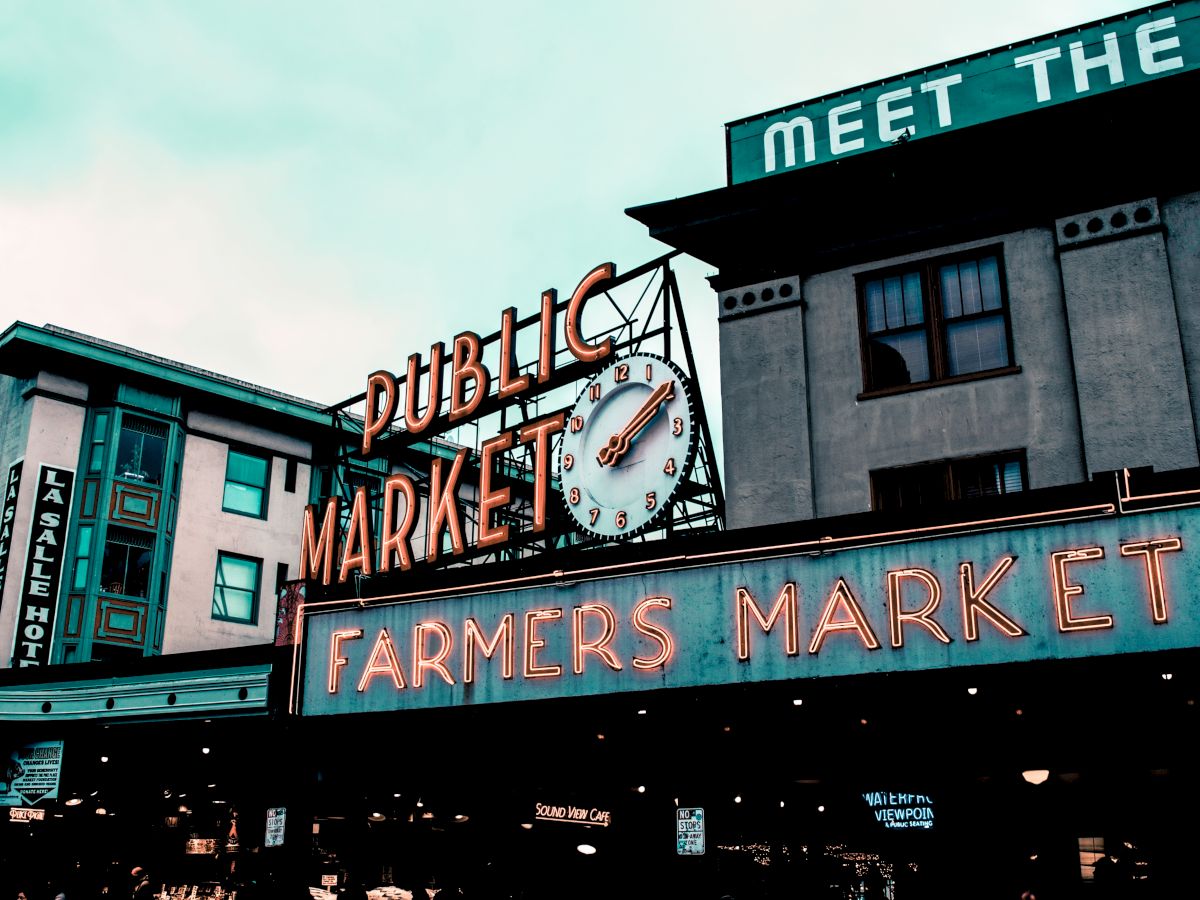 The image shows a neon sign reading "Public Market" and "Farmers Market" with a clock, against a backdrop of buildings under a cloudy sky.