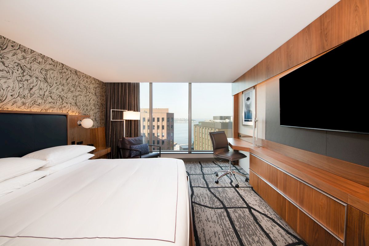 A modern hotel room with a large bed, desk, chair, TV, and a window view of buildings and water.