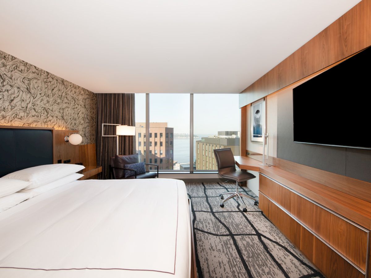 A modern hotel room with a large bed, desk, chair, TV, and a window view of buildings and water.