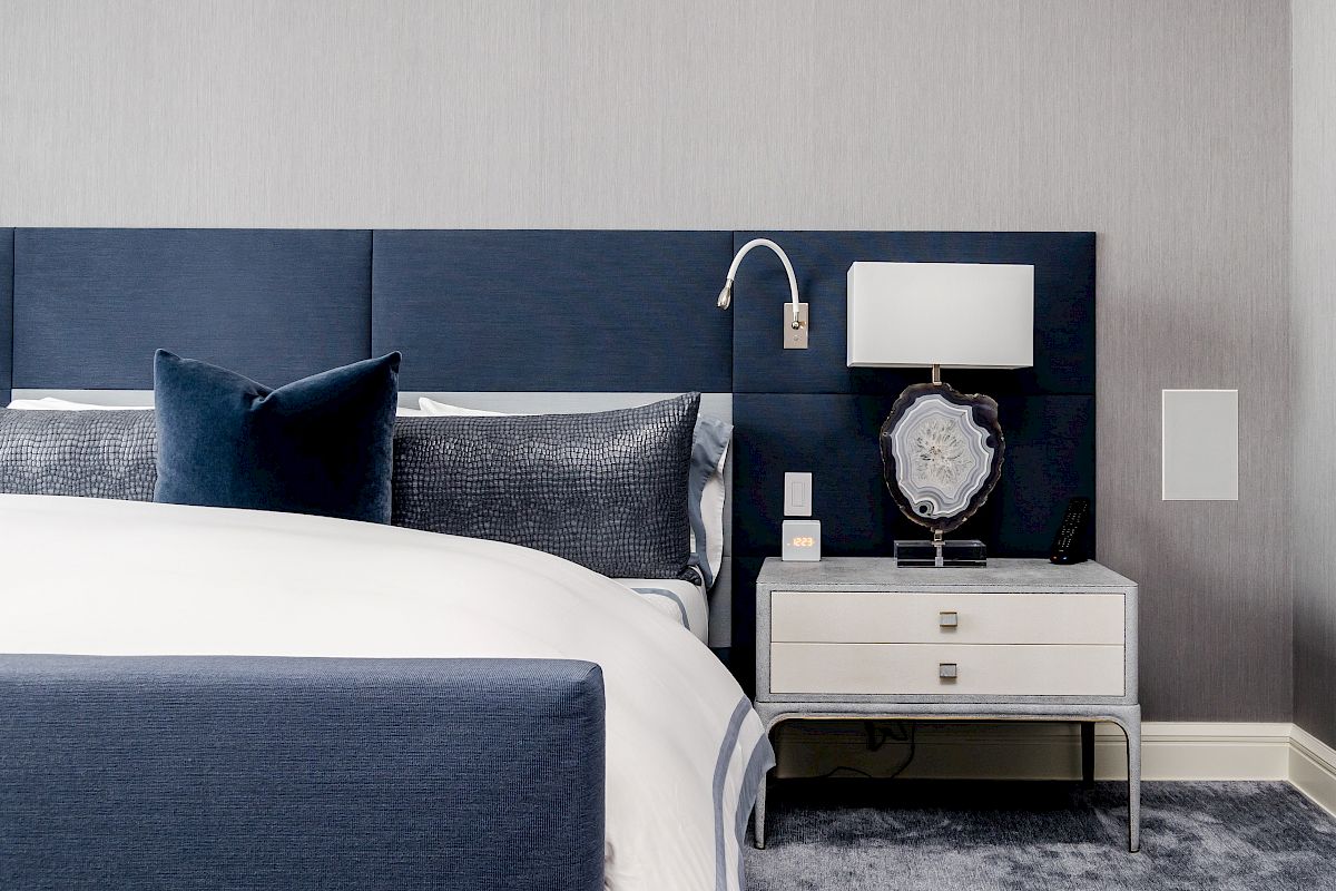 The image shows a modern bedroom with a blue headboard, pillows, a bed, a nightstand with a lamp, and a carpeted floor.