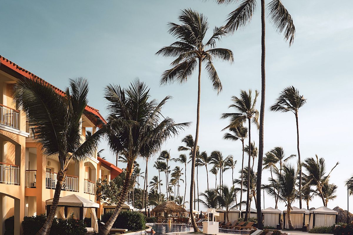 A tropical resort pool area with palm trees, lounge chairs, and a building under a clear sky.