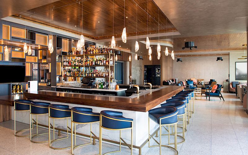 A modern bar with blue stools, hanging lights, and a well-stocked shelf of bottles, set in a stylish lounge area with seating.